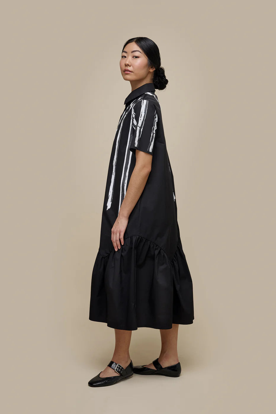 A woman with long dark hair is wearing the UCHUU LONG BLOUSE STYLE DRESS WITH SHORT SLEEVES, a black midi dress adorned with white vertical stripes and buttons down the front. The dress, designed to mimic a long blouse, features a ruffled hem. She stands against a plain beige background and pairs the outfit with black shoes.