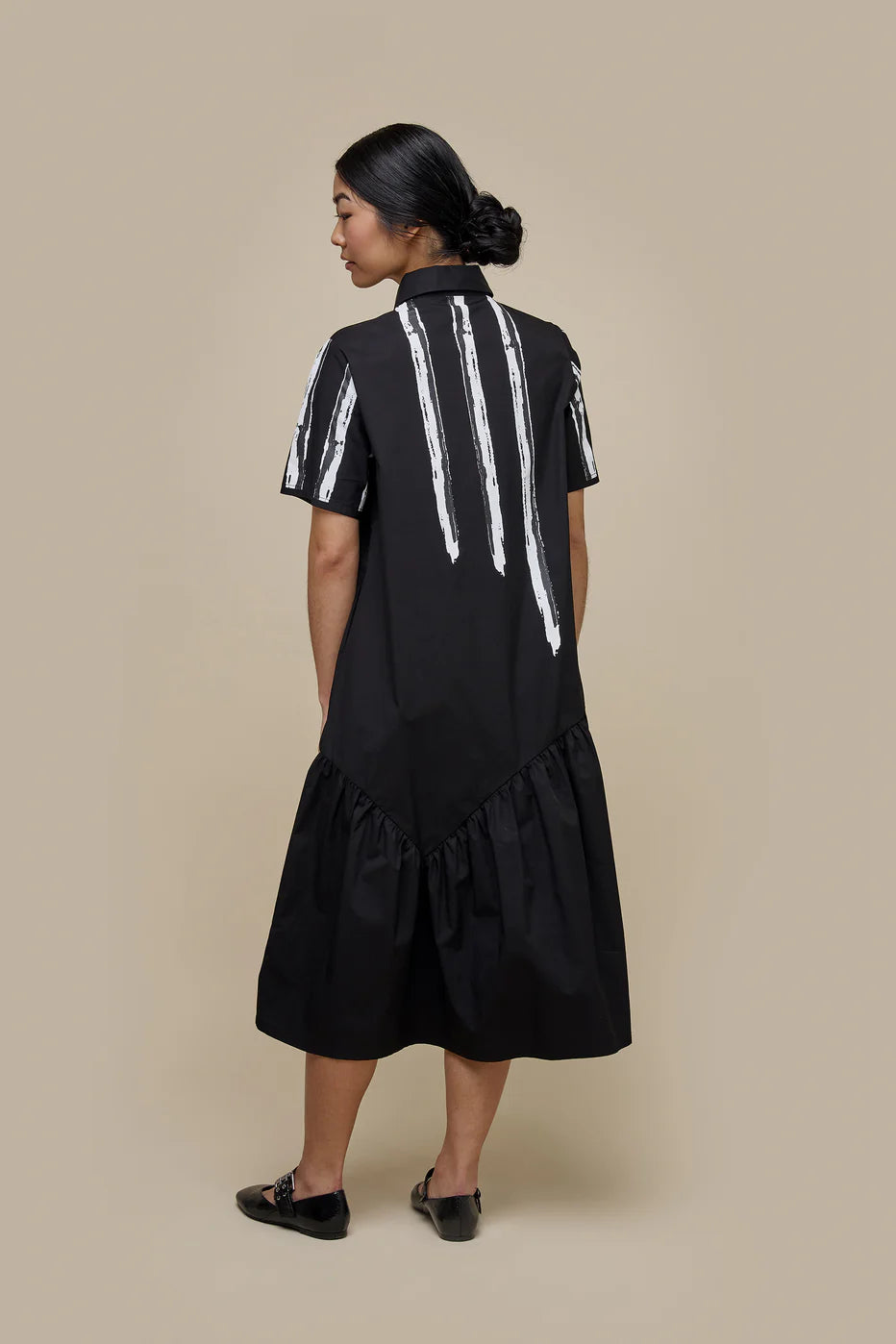 A woman with long dark hair is wearing the UCHUU LONG BLOUSE STYLE DRESS WITH SHORT SLEEVES, a black midi dress adorned with white vertical stripes and buttons down the front. The dress, designed to mimic a long blouse, features a ruffled hem. She stands against a plain beige background and pairs the outfit with black shoes.