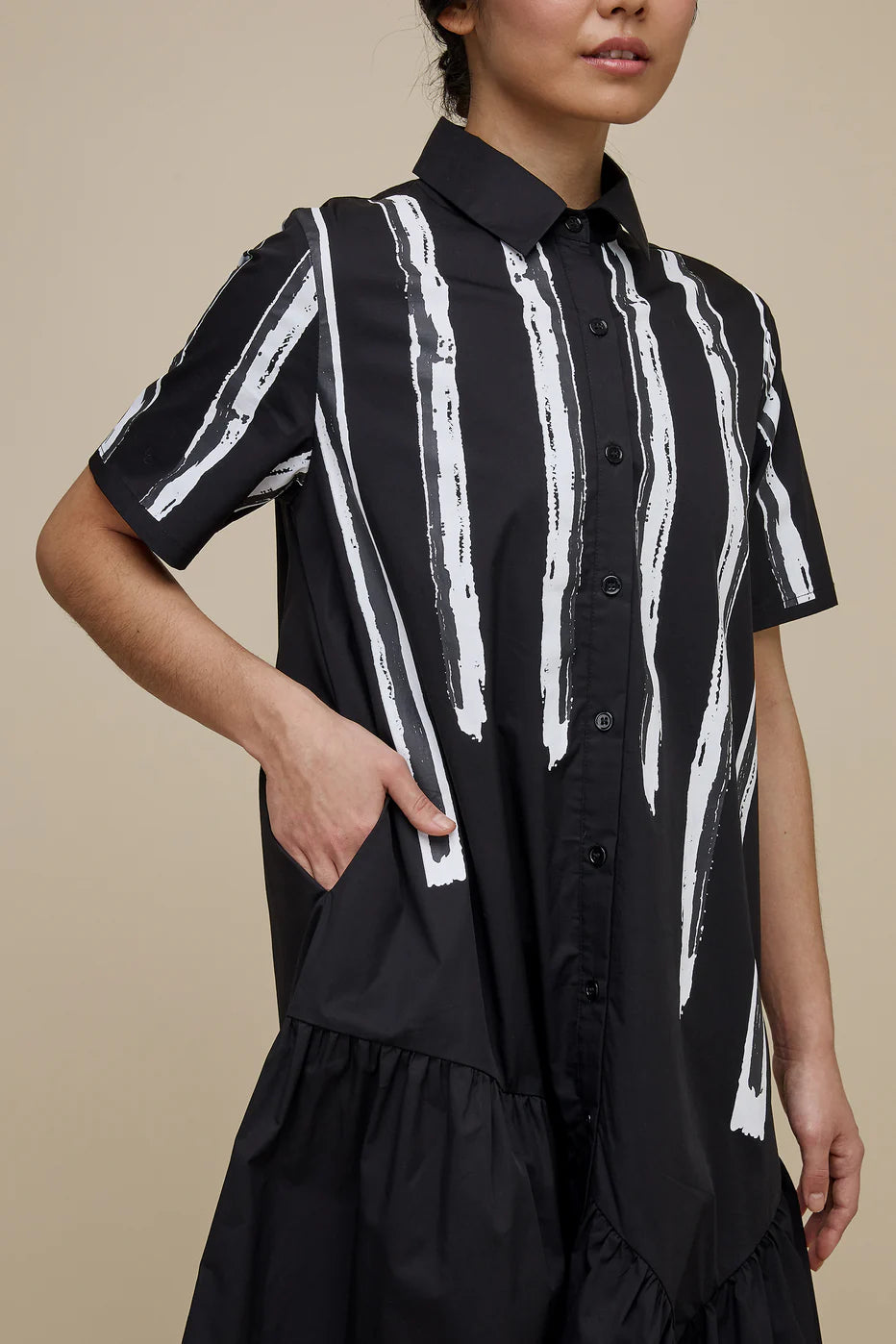A woman with long dark hair is wearing the UCHUU LONG BLOUSE STYLE DRESS WITH SHORT SLEEVES, a black midi dress adorned with white vertical stripes and buttons down the front. The dress, designed to mimic a long blouse, features a ruffled hem. She stands against a plain beige background and pairs the outfit with black shoes.