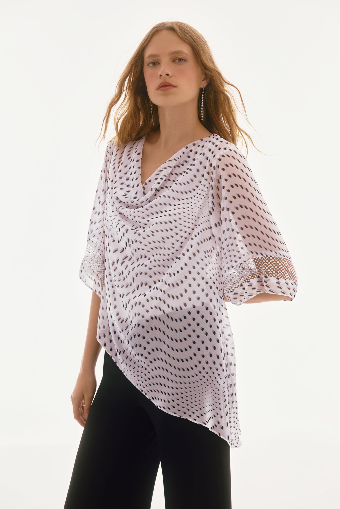 A woman stands confidently in the Signature Chiffon Dot Print Layered Top 251774 by Joseph Ribkoff, her loose, wavy hair cascading naturally. She pairs this chic white chiffon top with black dots and bell sleeves with black pants, showcasing her effortlessly stylish look.