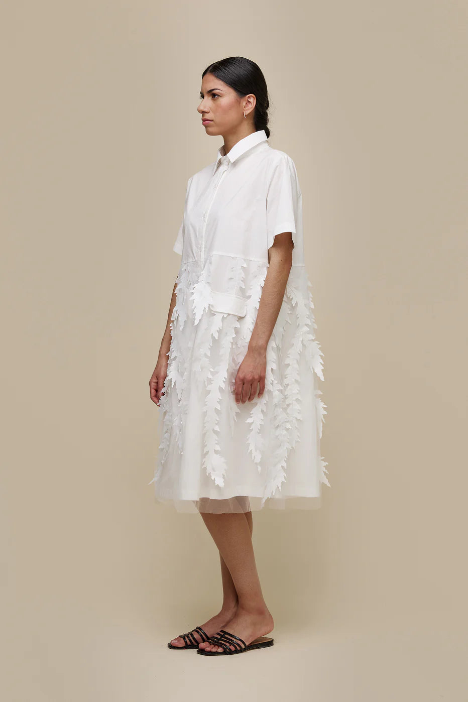 A person with long dark hair in a braid, dressed in a white, short-sleeved, knee-length UCHUU A-LINE BLOUSE STYLE DRESS with leaf-patterned organza appliqué embellishments and invisible side pockets (Product ID: CS24-009), stands against a plain beige background. They wear black slip-on sandals and have a neutral expression on their face.