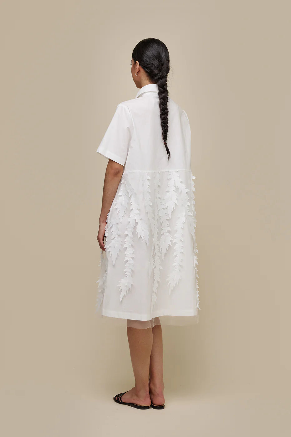A person with long dark hair in a braid, dressed in a white, short-sleeved, knee-length UCHUU A-LINE BLOUSE STYLE DRESS with leaf-patterned organza appliqué embellishments and invisible side pockets (Product ID: CS24-009), stands against a plain beige background. They wear black slip-on sandals and have a neutral expression on their face.
