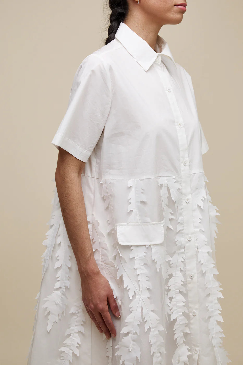 A person with long dark hair in a braid, dressed in a white, short-sleeved, knee-length UCHUU A-LINE BLOUSE STYLE DRESS with leaf-patterned organza appliqué embellishments and invisible side pockets (Product ID: CS24-009), stands against a plain beige background. They wear black slip-on sandals and have a neutral expression on their face.