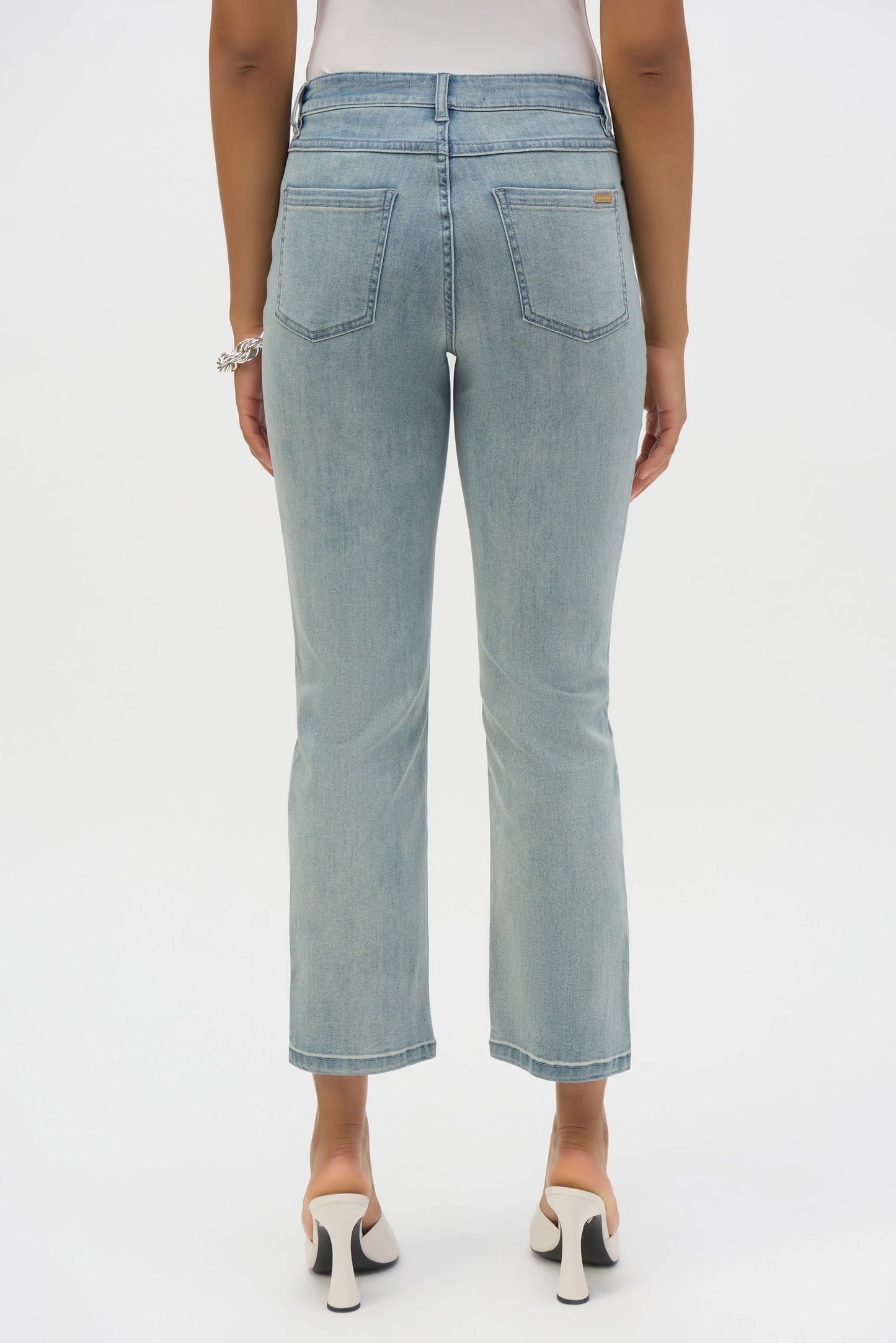 Rear view of a person in Joseph Ribkoff's Margot Classic Straight Leg Stretch Jeans, crafted from Featherweave denim, paired with white heeled shoes. The light blue jeans have two back pockets and a vintage wash charm. A bracelet graces their right wrist against a plain white backdrop.