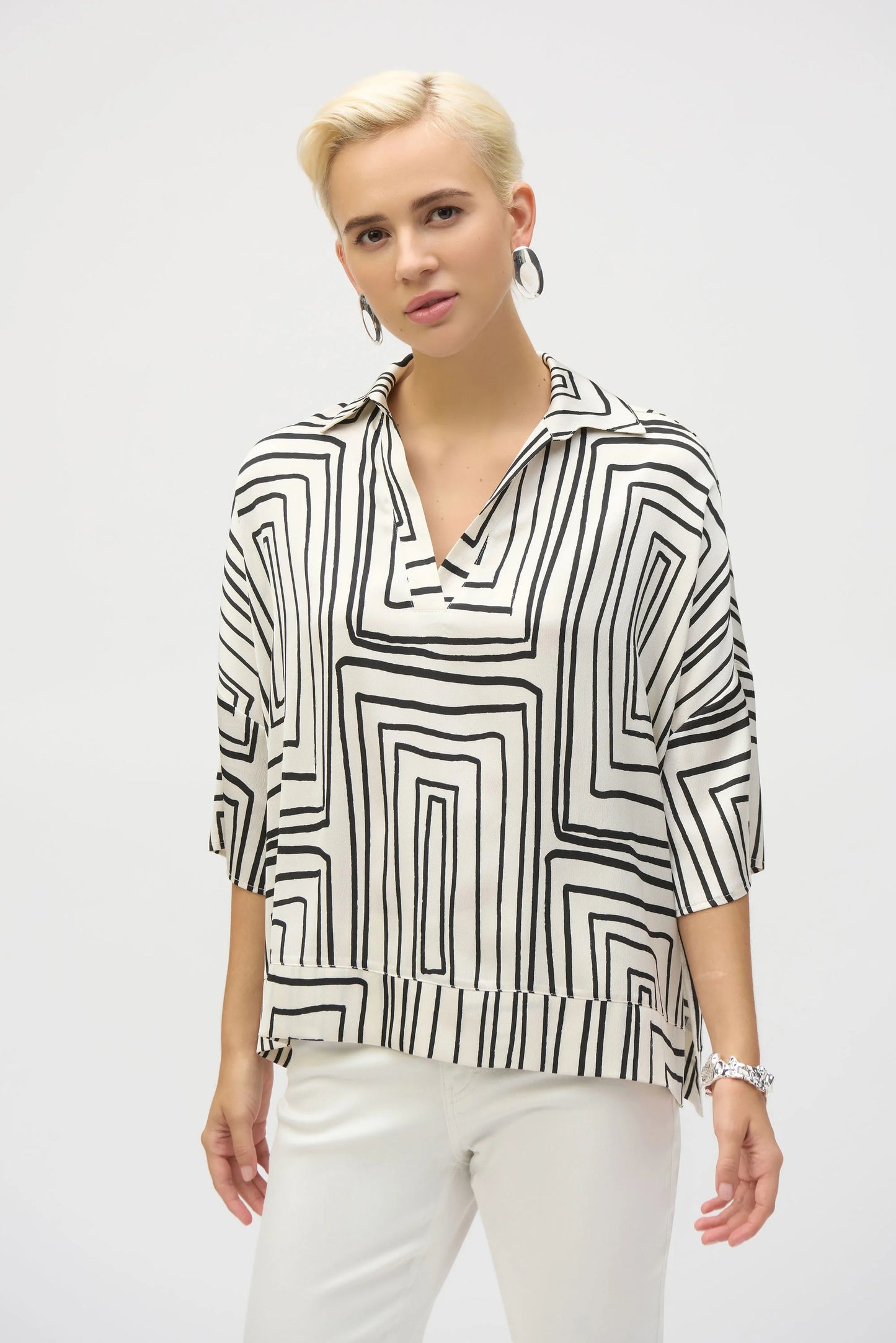 A person with short blonde hair wears a Joseph Ribkoff Satin Geometric Print Boxy Top 252060 and white pants, accessorized with hoop earrings and a bracelet, while looking into the camera against a plain background.