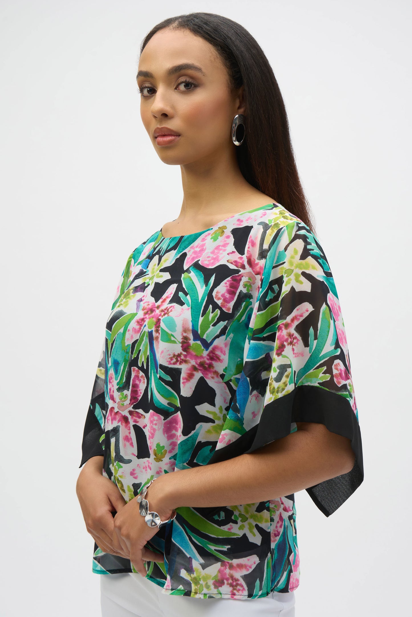 A woman with long dark hair models the Joseph Ribkoff Chiffon Tropical Print Straight Top 252126 and white pants against a plain white background. She complements her look with silver hoop earrings and a bracelet, gazing confidently at the camera.