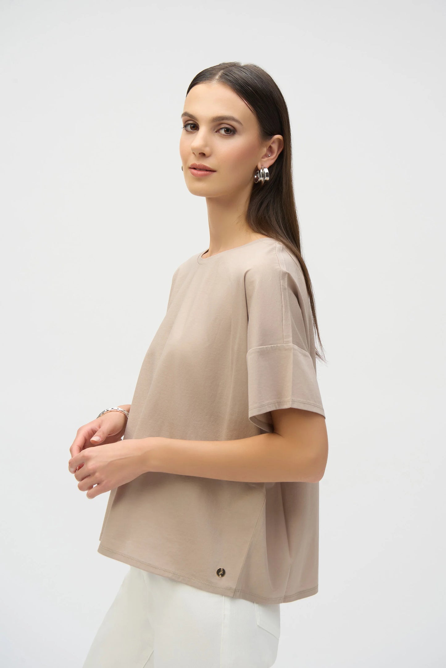 A person with long brown hair wears a Joseph Ribkoff Organic Cotton Boxy T-Shirt in beige and white pants. Standing against a plain white background, they have a neutral expression and glance slightly over their shoulder.