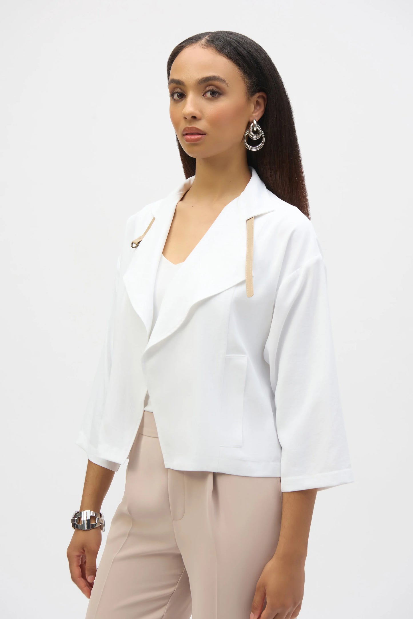 A woman with long, dark hair stands against a plain background, wearing a Joseph Ribkoff Eggshell Boxy Cover-Up 252149 blazer. The loose-fit pairs with a light top, beige trousers, and silver hoop earrings. She gazes slightly to the side with a calm expression.