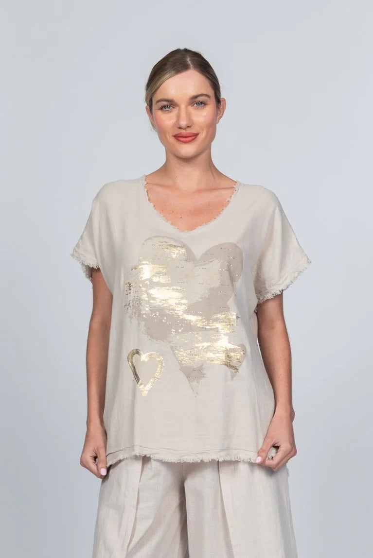 The woman wears a light beige Linen Blend T-shirt MCK2502B/A by Paolo Tricot, adorned with a large gold heart at the center. Made in Italy, this breathable top enhances her styled-back hair and slight smile against the plain backdrop.
