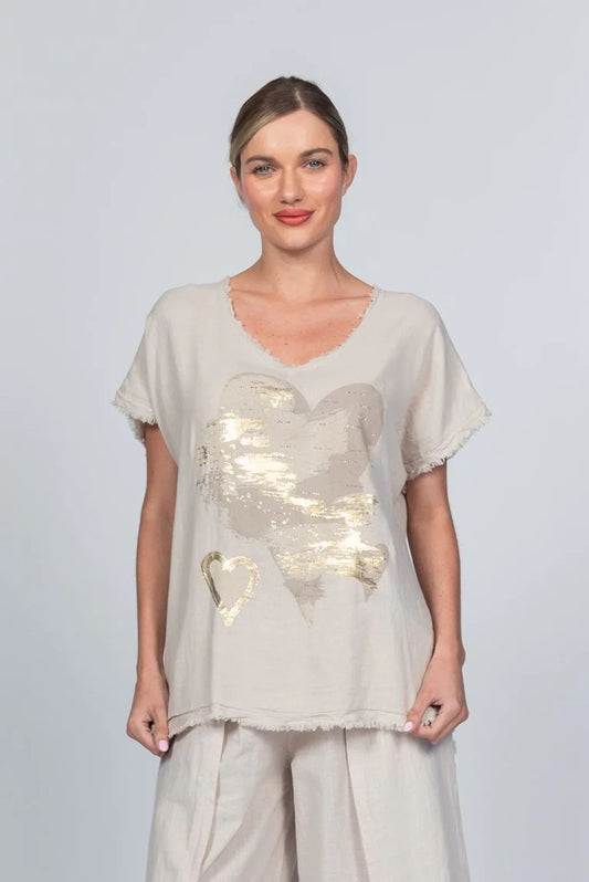 The woman wears a light beige Linen Blend T-shirt MCK2502B/A by Paolo Tricot, adorned with a large gold heart at the center. Made in Italy, this breathable top enhances her styled-back hair and slight smile against the plain backdrop.