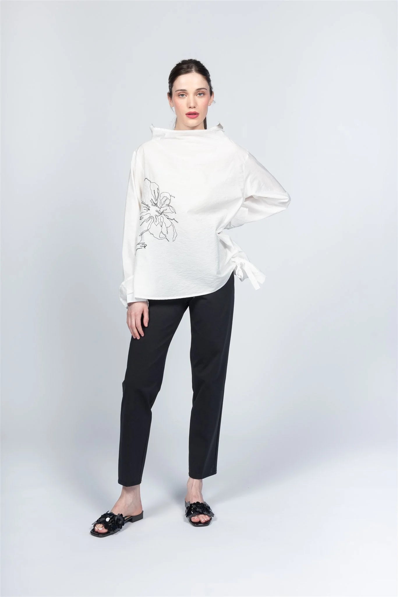 Against a light gray background, a person wears the elegant Italian-crafted Paolo Tricot blouse (Top WT230242 - Pablo Tricot) in O.WHITE with floral embroidery and black pants. Their hair is tied back, black sandals adorn their feet, one hand rests on their hip while the other hangs by their side.