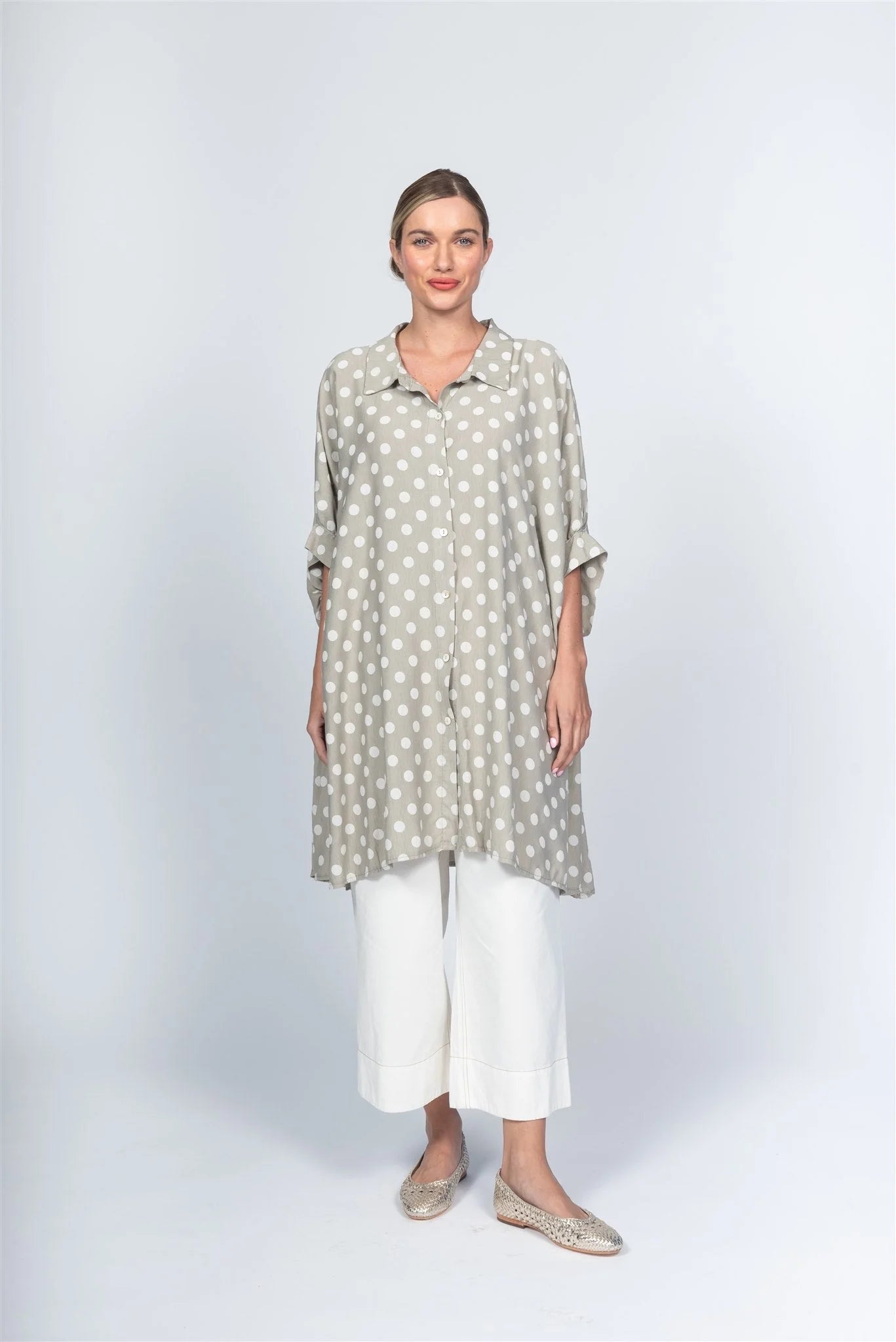 Against a plain backdrop, a person dons a loose gray polka dot tunic with white pants and casual taupe slip-ons by Paolo Tricot, exuding effortless Italian style.