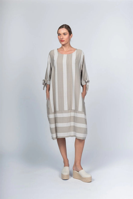 The woman wears the Dress DM80112 Taupe by Paolo Tricot, a loose-fitting, knee-length piece with taupe and white stripes, tie-detail sleeves, and made from 100% viscose. It’s paired with light platform shoes against a plain light gray backdrop.