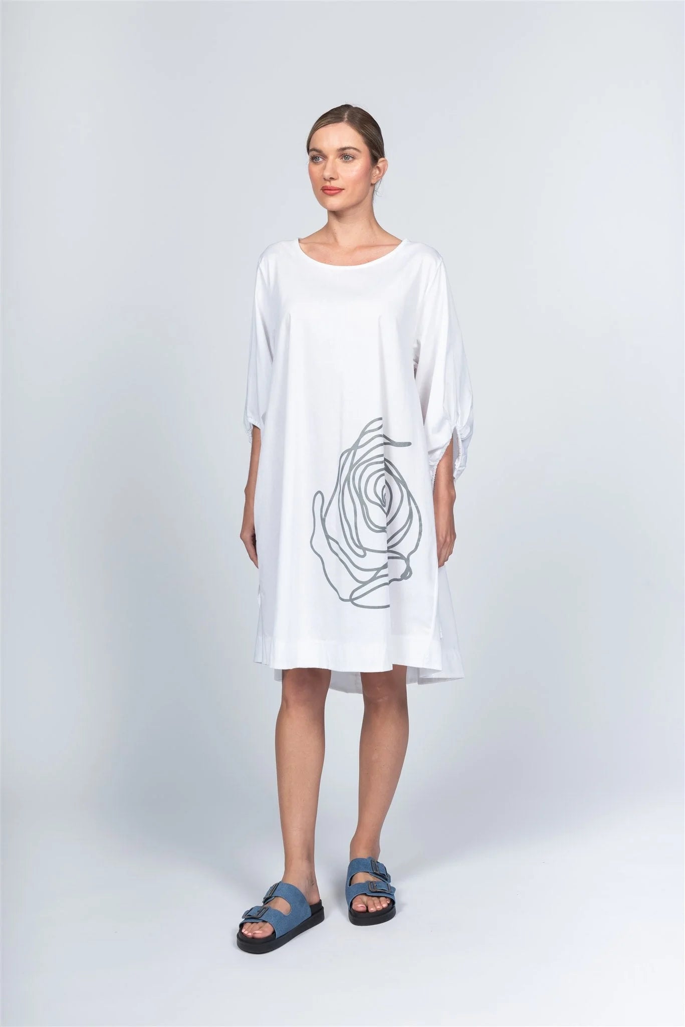 Against a grey backdrop, a woman wears the loose Dress AL5196 by Paolo Tricot, featuring an abstract black rose design. Her hair is tied back and she completes the look with blue sandals. Made in Italy, this piece is a timeless wardrobe addition.