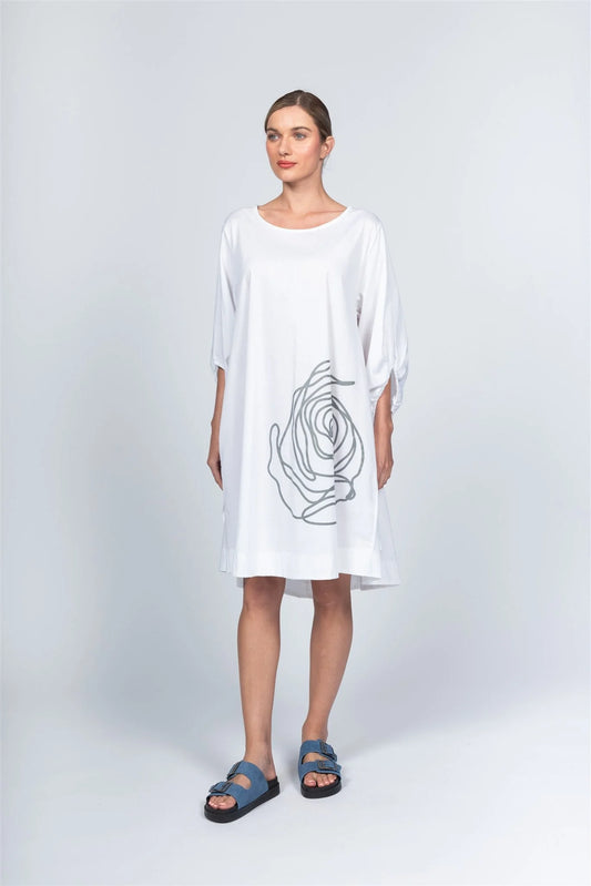 Against a grey backdrop, a woman wears the loose Dress AL5196 by Paolo Tricot, featuring an abstract black rose design. Her hair is tied back and she completes the look with blue sandals. Made in Italy, this piece is a timeless wardrobe addition.