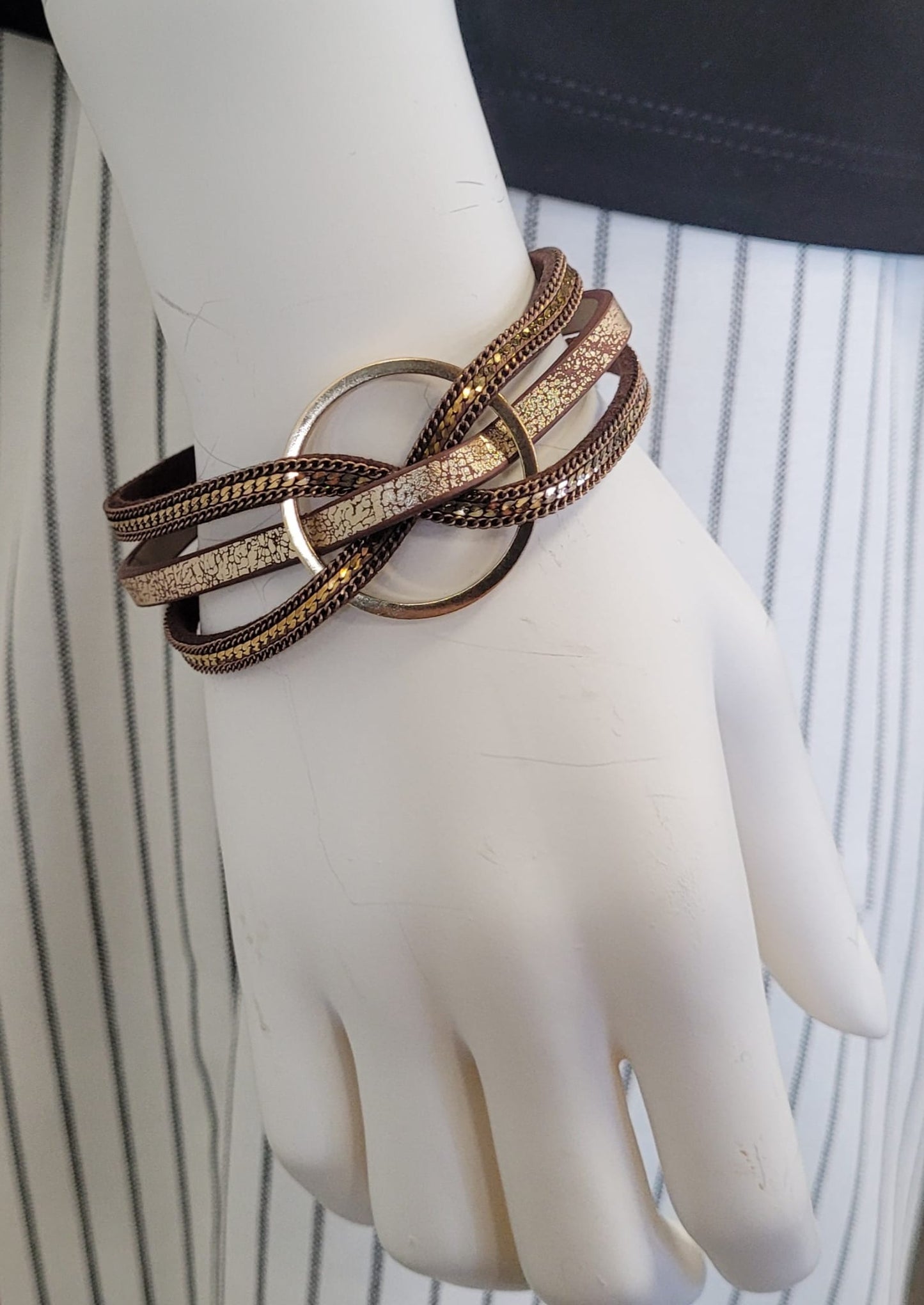 A mannequin hand displays the Clara 15 Bracelet by Elise accessories, with multiple strands looping through a central gold ring, set against black and white vertical stripes—a magnetic piece that adds modern flair to any outfit.