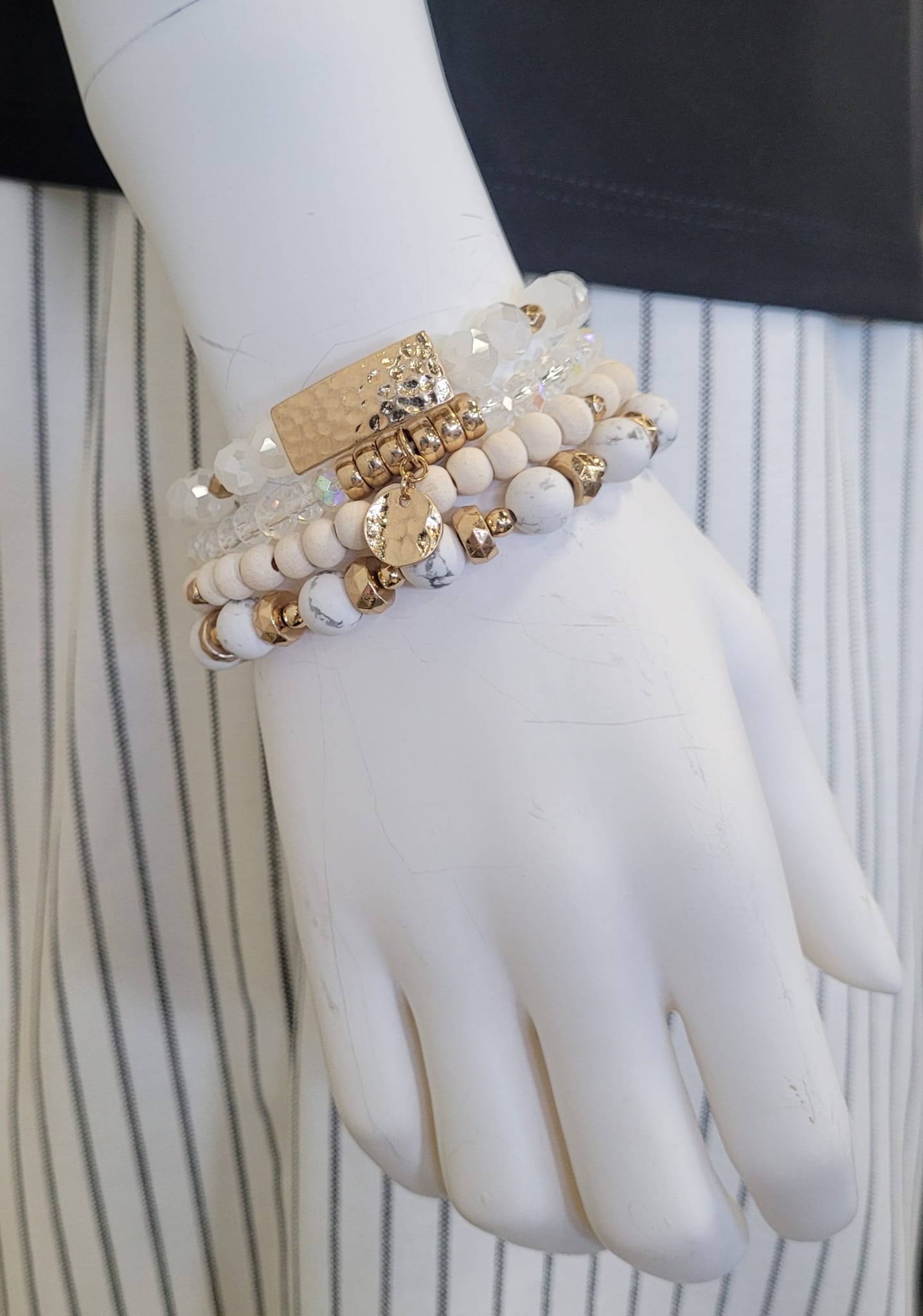 A mannequin hand displays the Gina 26 Bracelet by Elise accessories, a chic piece with layered white, beige, gold, and clear beads, shown with a black top and white skirt with black stripes.