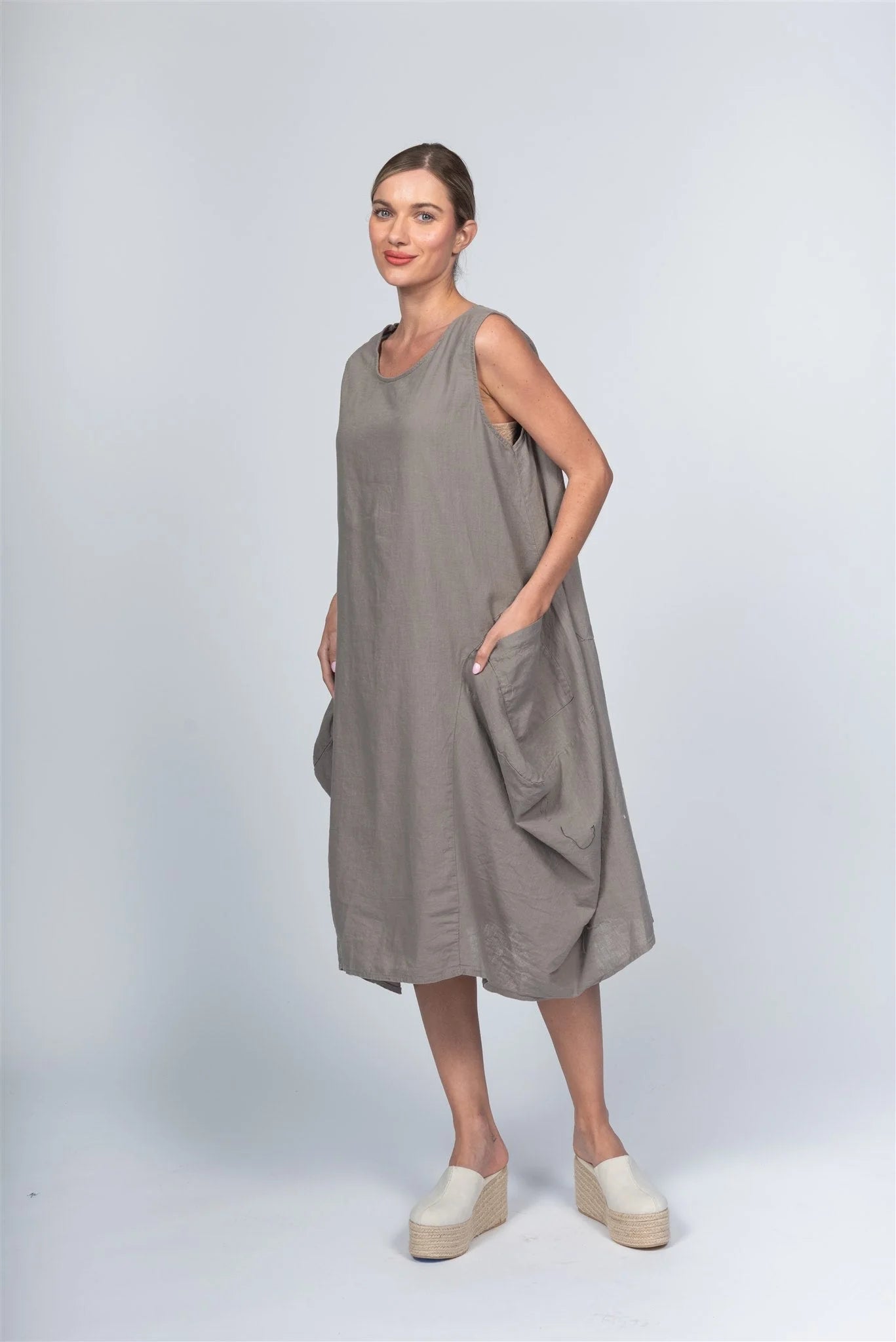 A woman stands on a light background wearing a loose, sleeveless gray Dress Linen Blend by Paolo Tricot. She has her left hand in the side pocket, paired with white platform shoes. Her hair is tied back as she smiles slightly, embodying luxury and comfort with PAOLO TRICOT elegance.
