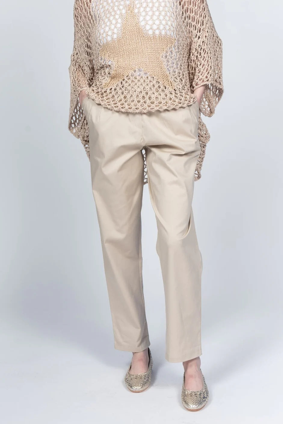 A person wearing Pablo Tricot Pants with Belt LX38551 and a beige mesh sweater featuring a large star design enjoys the comfort of their gold loafers. Made in Italy, this chic ensemble stands out against the light gray background, with hands casually resting in pockets.