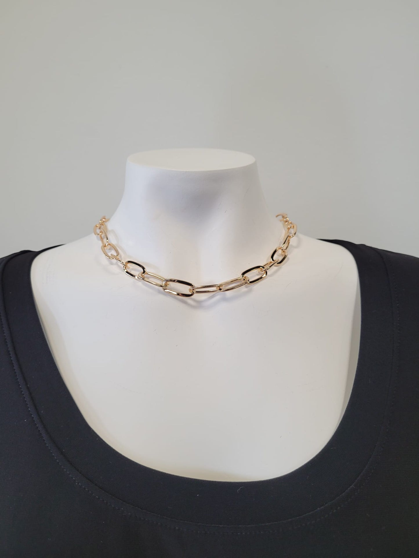 The Rhea 4 Necklace by Elise Accessories, featuring large gold oval links, is showcased on a white mannequin dressed in a black round-neck top against a light plain background.