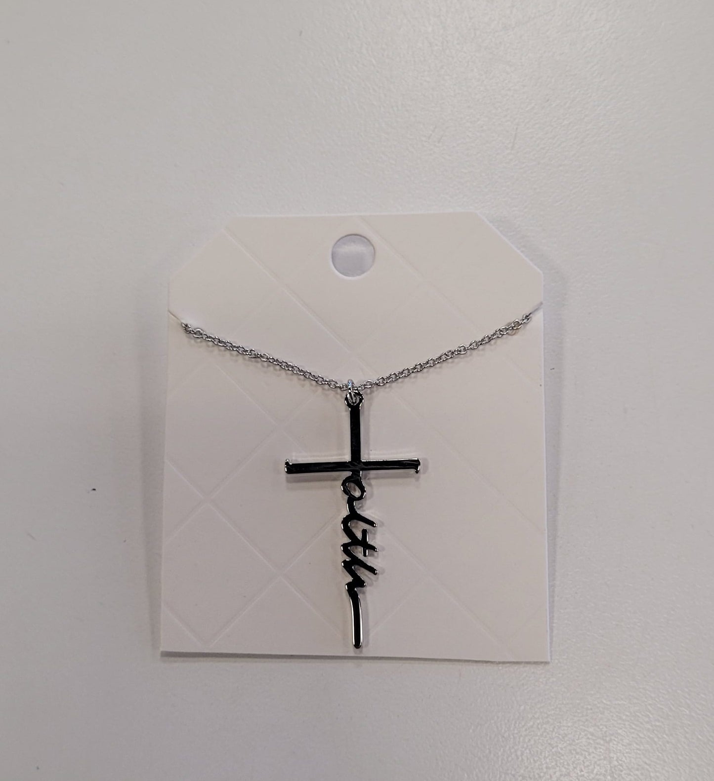 The Trina 13 Necklace by Elise accessories features "faith" woven into a silver cross, displayed on a diamond-patterned white card—a stylish addition to any collection.