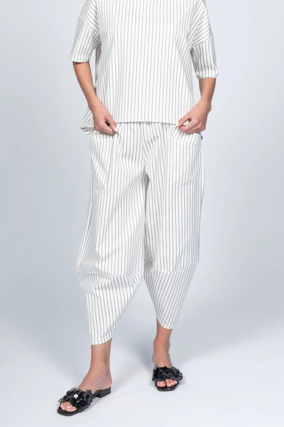 Wearing a stylish white outfit with black vertical stripes, including Pants Striped 33760 by Paolo Tricot and a short-sleeved top, a person casually rests their hands in pockets. Their look is completed by comfy black slide sandals adorned with flowers, all set against a plain light gray backdrop.