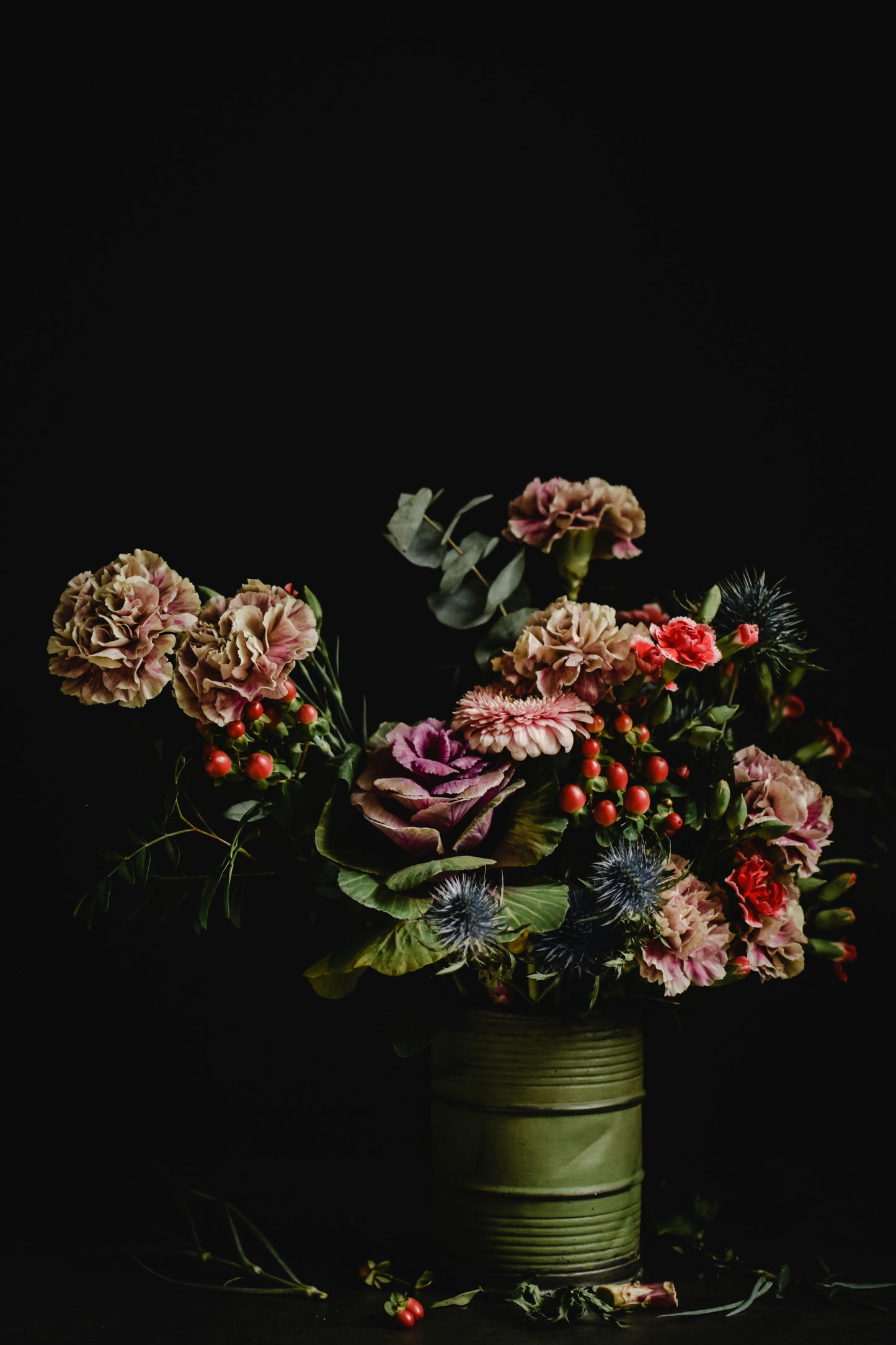 The Designer’s Choice Centrepiece by Posies Flowers & Fashion features a clear glass vase filled with a vibrant bouquet of assorted flowers, including pink roses, peach roses, blue delphiniums, and small yellow daisies. This exquisite flower arrangement is showcased on a table in the foreground of a softly lit room with blurred furniture and greenery in the background.