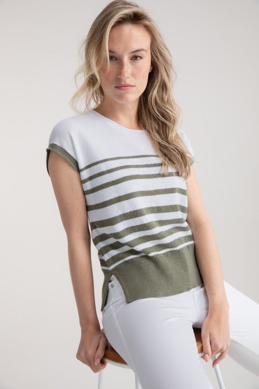 A person with long blonde hair sits on a stool, wearing an elegant and comfortable MARBLE Sweater Top 7881 in white and green stripes, made from 100% cotton, paired with white pants. The background is plain and light.