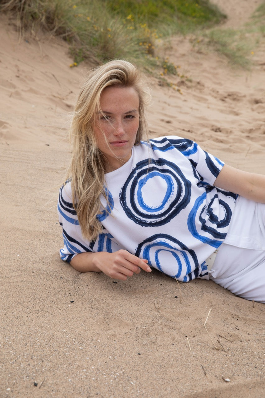 On a sandy beach, a person with long blonde hair relaxes, dressed in MARBLE's Sweater Top 7809 MARBLE and white pants, creating a luxurious vibe. The scene is set against grass and sparse vegetation.