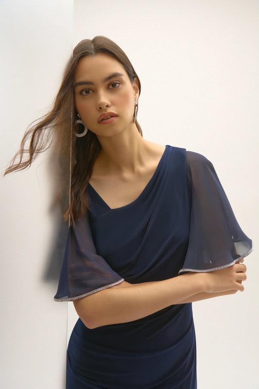 A woman poses against a white wall in the Signature Silky Knit and Chiffon Sheath Dress by Joseph Ribkoff. With butterfly sleeves and an asymmetrical neckline, she wears hoop earrings and gazes softly, her long hair gently moved by a breeze.