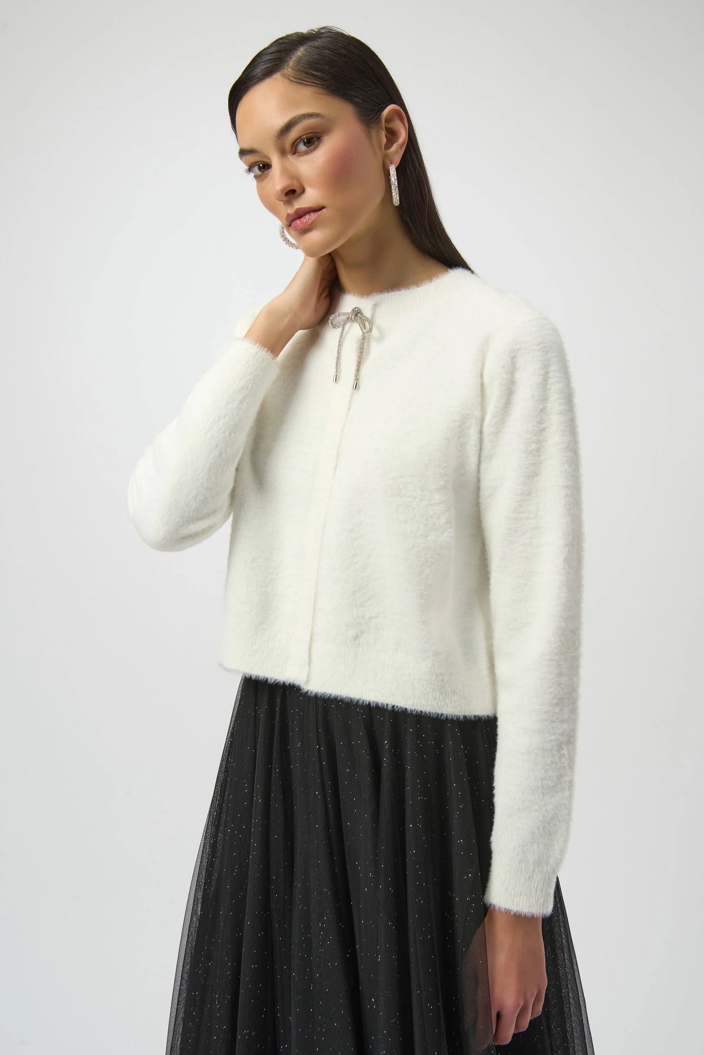 A woman with straight dark hair models the Joseph Ribkoff Feather Yarn Cardigan with Rhinestone Bow 254955 against a plain light background, gazing softly at the camera.