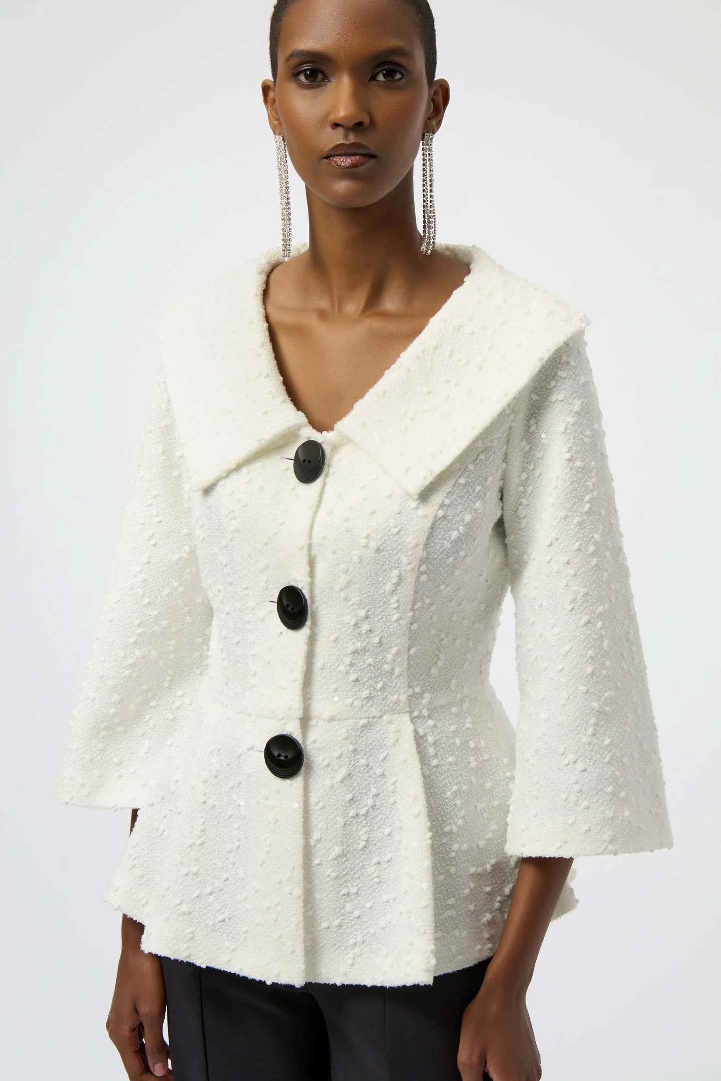 A person wears the Joseph Ribkoff Sequined Bouclé Knit Peplum Jacket 254184—textured white with large black buttons, wide collar, and three-quarter bell sleeves—paired with dark pants against a plain background.