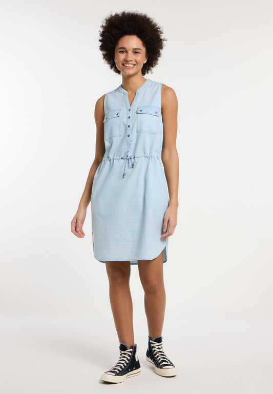 A person with curly hair smiles, wearing a ROISSIN DENIM DRESS 2511-20034-2015 by RAGWEAR, a light blue sleeveless relaxed fit dress with a Henley neckline and buttoned chest pockets. They pair it with black high-top sneakers against a plain white background.