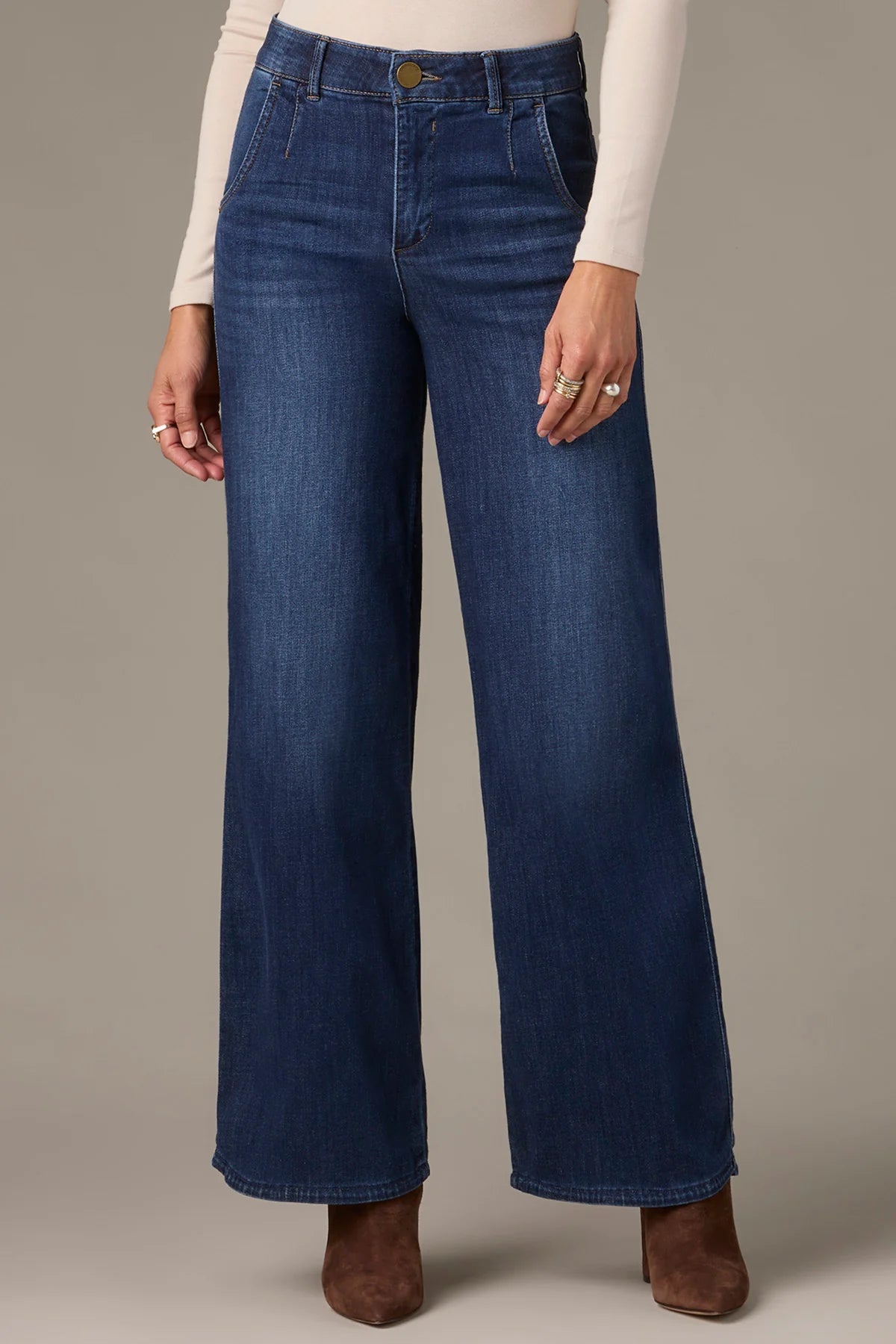 A person wearing Wit & Wisdom WHITNEY SKYRISE WIDE LEG JEANS in blue, paired with a cream long-sleeve top and brown heeled boots, stands against a plain background.