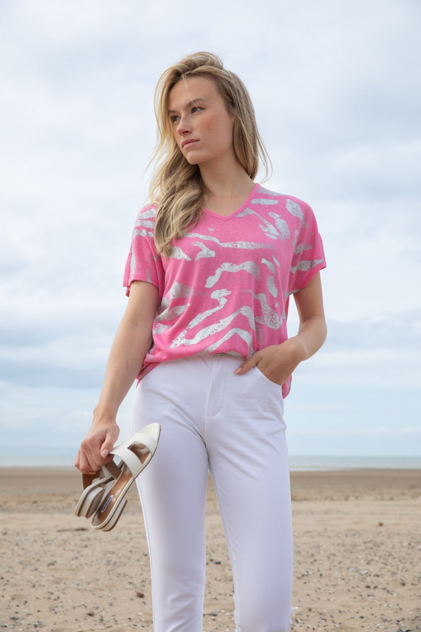 On the beach, a person clutches their sandals and wears Top 7371 by MARBLE with white pants, as a cloudy sky casts shadows on the sand.