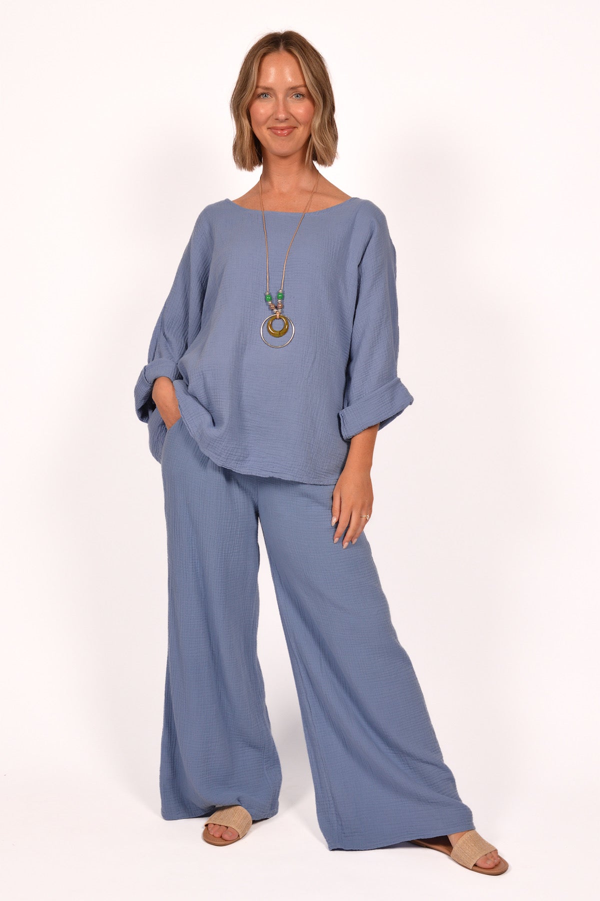 A person is wearing the Cotton Gauze Pants ITS70031 and a matching top by Catherine Lilywhite. The blue outfit, made in Italy, is loose-fitting. They accessorize with a long necklace with circular pendants and sandals, posing with one hand in a pocket against a plain white background.