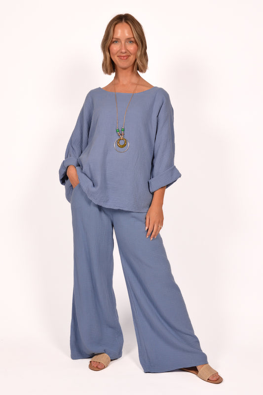A person is wearing the Cotton Gauze Pants ITS70031 and a matching top by Catherine Lilywhite. The blue outfit, made in Italy, is loose-fitting. They accessorize with a long necklace with circular pendants and sandals, posing with one hand in a pocket against a plain white background.