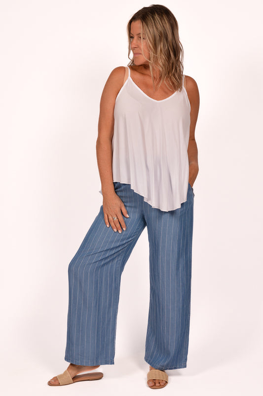 A woman poses in Catherine Lilywhite's blue striped Tencel Pant ITIM23163BJ and a white sleeveless top, showcasing Italian craftsmanship. She wears brown sandals, with shoulder-length hair turned to the side against a plain summer backdrop.