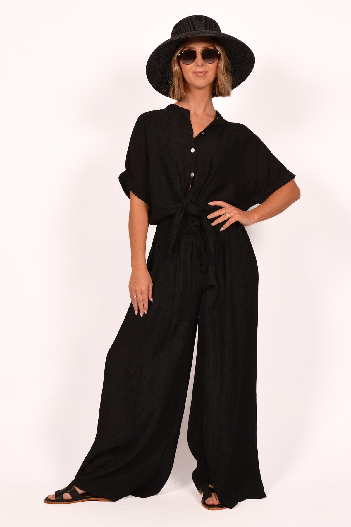 A person poses confidently in a Catherine Lillywhite Italian-made Black Textured Tie Front Blouse ITEL807581BK and wide-legged pants, wearing a wide-brimmed hat and sunglasses. Paired with relaxed sandals, this creates an effortlessly chic look against a plain background.