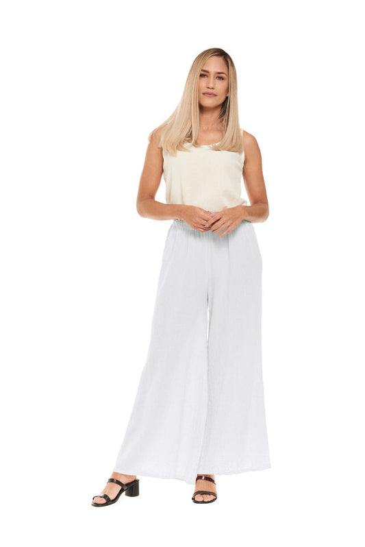 A person with long hair wearing a sleeveless top and Oh My Gauze's SURF SNOW PANT confidently stands. The wide-leg cotton gauze pants offer a relaxed waist for comfort. Clasping their hands in front, they wear heeled sandals against a plain white backdrop.