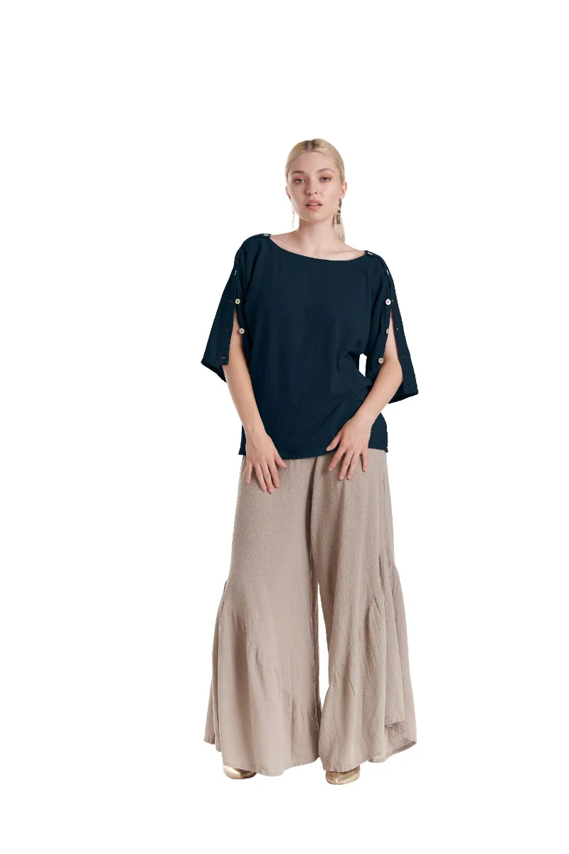 A person wearing the SAINT SAPPHIRE TOP by Oh My Gauze, a navy cotton gauze top with buttoned sleeves, pairs it with loose beige wide-leg pants. Against a plain white background, their light-colored hair is tied back, crafting a stylish and classic look that exudes casual comfort.