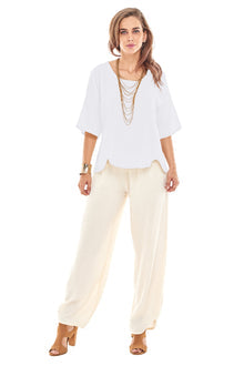 A person stands wearing the Oh My Gauze SCALLOP SNOW TOP, cream wide-legged pants, and tan high heels. Accessories include layered necklaces and bracelets. The individual has long, wavy hair and poses with a relaxed, confident expression.