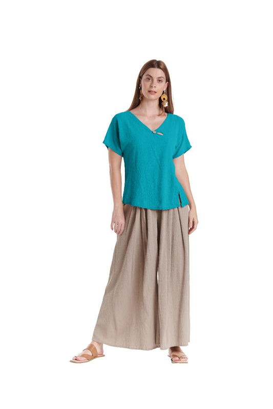 Against a white backdrop, a person wears the MIKE GLACIER TOP by Oh My Gauze in turquoise and beige wide-leg pants with front slits. Sandals and hoop earrings accessorize the look, while their long hair adds elegance.