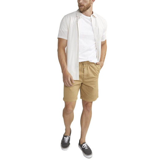 Wearing a white shirt over a t-shirt, the individual sports Silver Jeans Co.'s Pull-On Chino Short M52155CBT639, complemented by black sneakers with white soles. They stand against a plain white background with their left hand in their pocket.