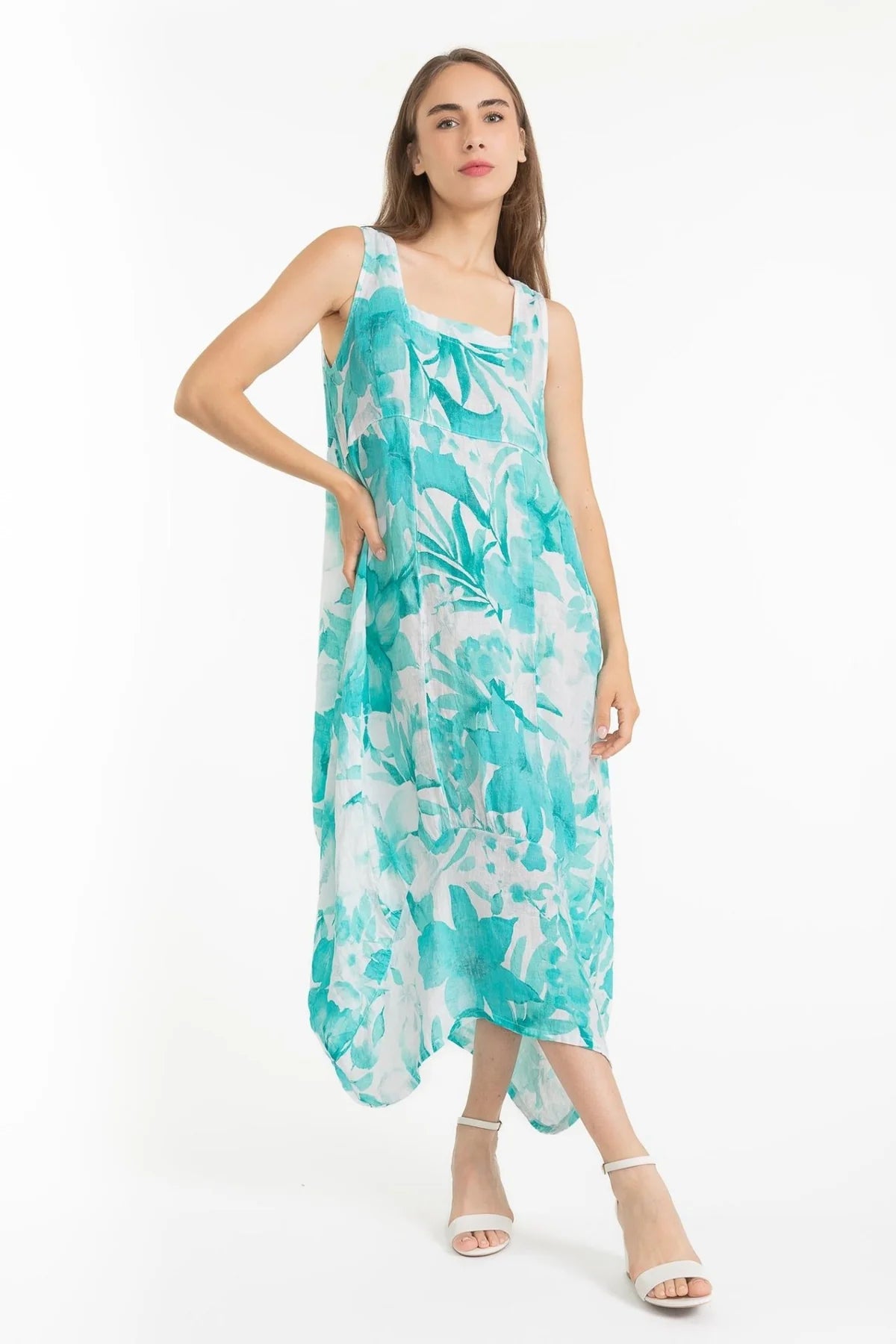 A woman models the French Linen Dress TP1612-11 Bianco from LinenLuv, its eco-friendly linen in a teal and white abstract pattern. In a white studio, she stands poised with one hand on her hip, her style enhanced by chic white strappy heels.