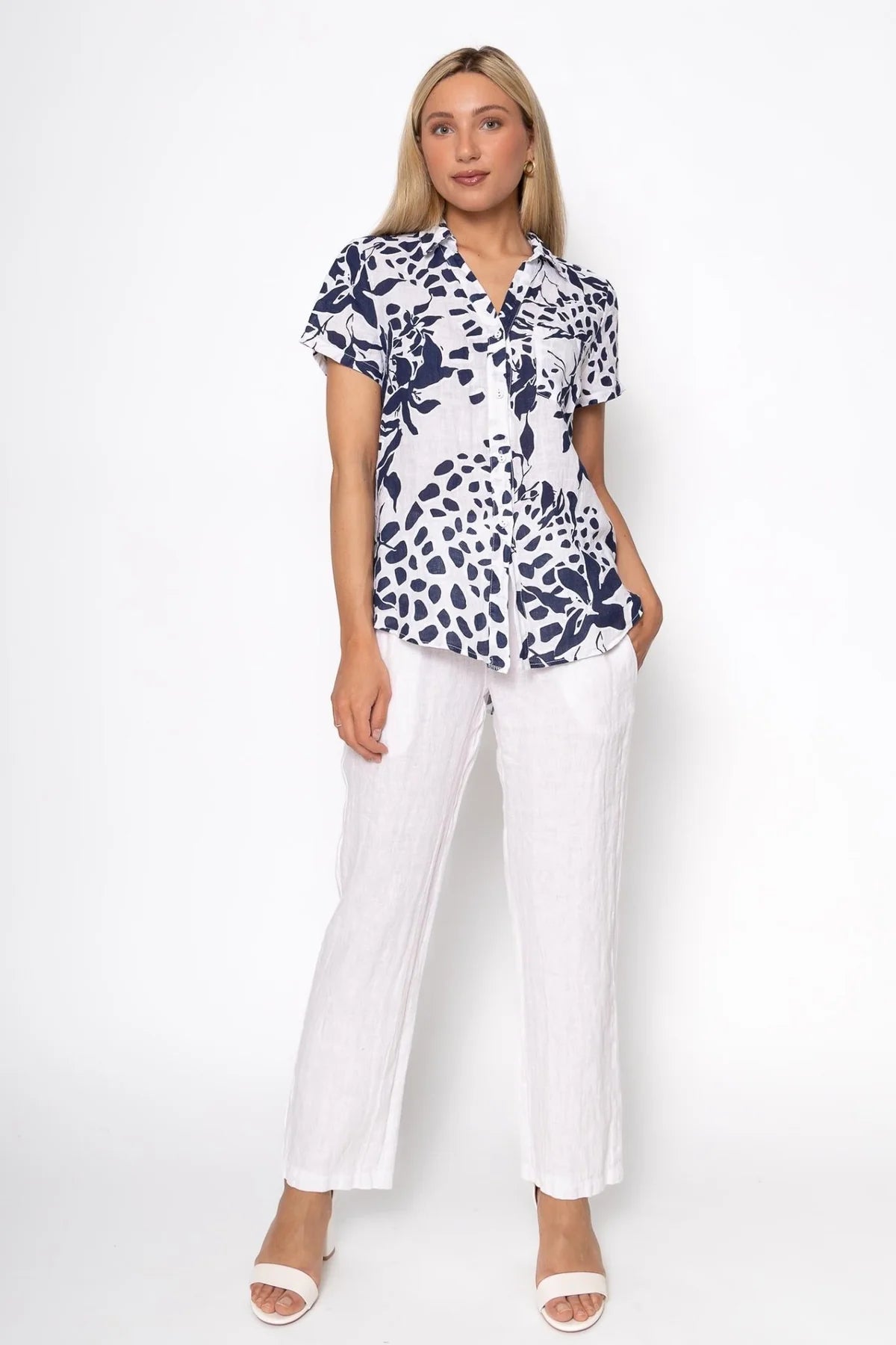 The individual is donning a French Linen Blouse TP1485-11 Bianco by LinenLuv, showcasing a navy and white floral pattern on eco-friendly fabric. Paired with white pants and sandals, they have long blonde hair against a plain white background.