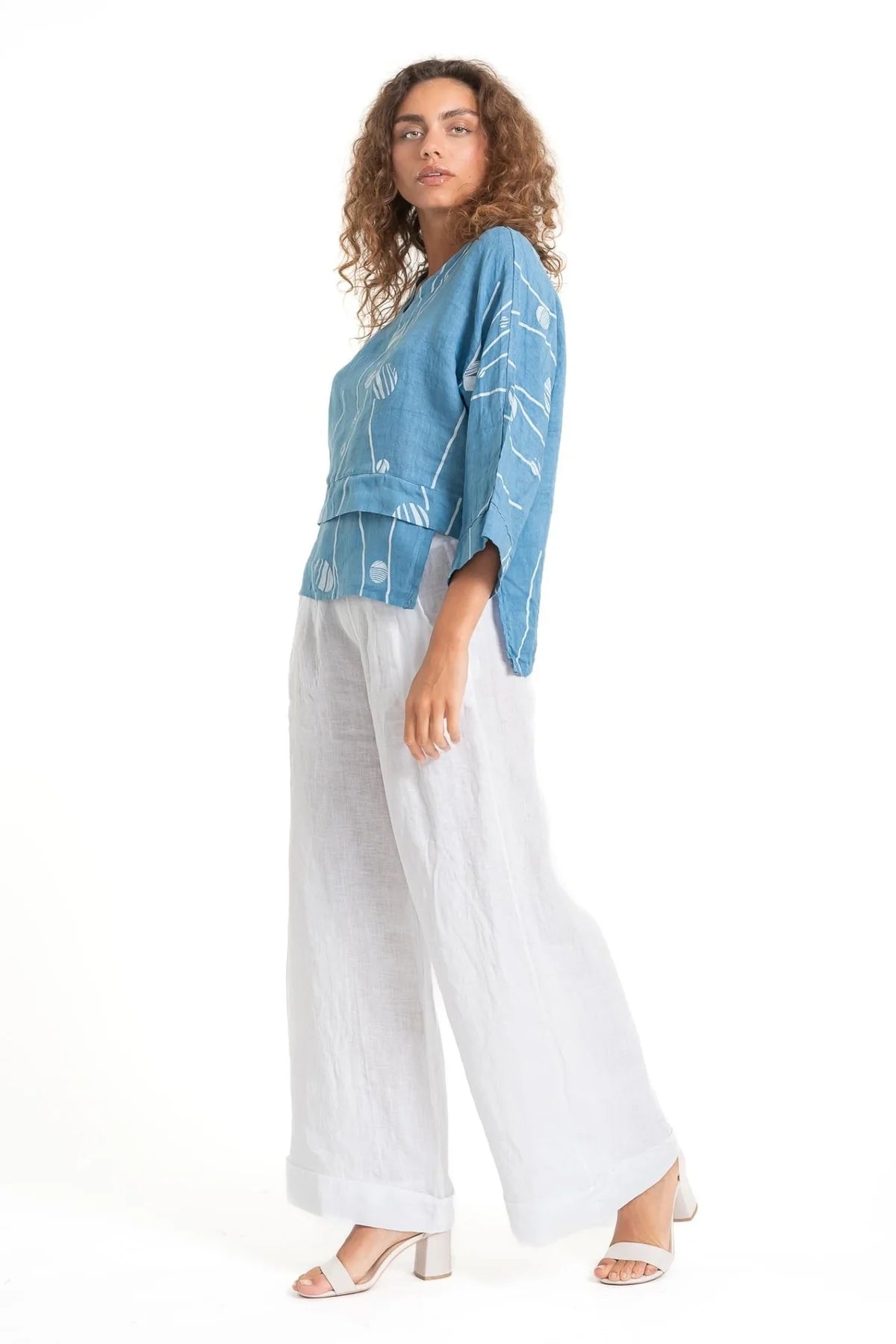 A curly-haired person wears the eco-friendly LinenLuv "French Linen Top TP1552P-185 Avio," featuring a blue shade with white patterns. They pair it with loose-fitting white pants and white heeled sandals, posing side-on against a plain white background.
