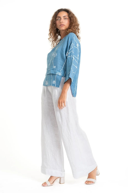 A curly-haired person wears the eco-friendly LinenLuv "French Linen Top TP1552P-185 Avio," featuring a blue shade with white patterns. They pair it with loose-fitting white pants and white heeled sandals, posing side-on against a plain white background.
