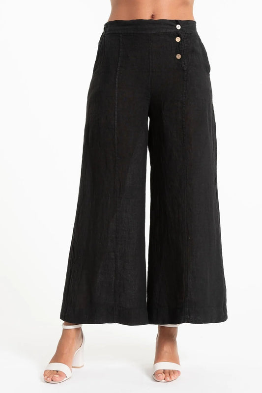 A person is partially visible, standing against a plain white background, wearing LinenLuv's French Linen Pant PT850-74 Nero with three side buttons and wide legs. They're paired with white open-toe sandals.