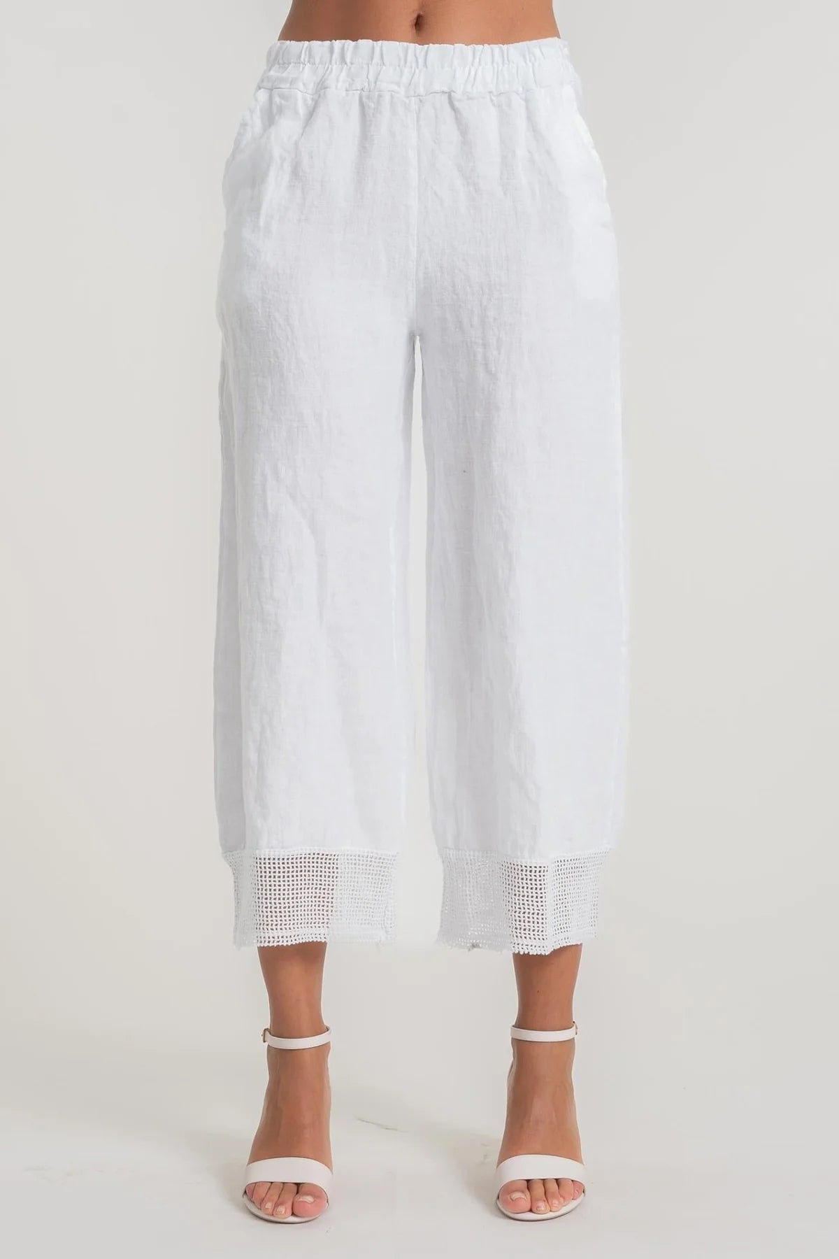 A person wearing LinenLuv's French Linen Pant PT840-11 Bianco, showcasing eco-friendly white linen with crochet cuff details, stands against a neutral background. The pants feature an elastic waistband and are paired with white open-toed heels.
